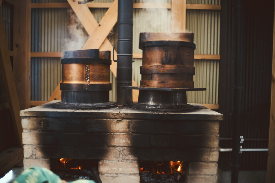 写真：かまどに置かれた2台の羽釜、蒸籠から湯気が吹き出している
